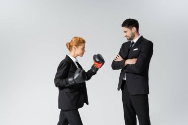 smiling businessman standing with crossed arms and looking at redhead businesswoman in boxing gloves isolated on grey clipart