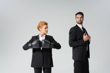 redhead businesswoman in boxing gloves looking at haughty businessman in stylish blazer isolated on grey clipart