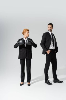 haughty businessman with hands in pockets looking at determined businesswoman in boxing gloves on grey background clipart