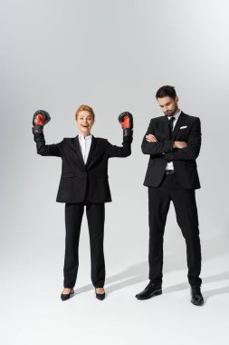 Boks eldivenli kızıl saçlı heyecanlı iş kadını gri arka planda üzgün iş adamının yanında zafer jesti sergiliyor.