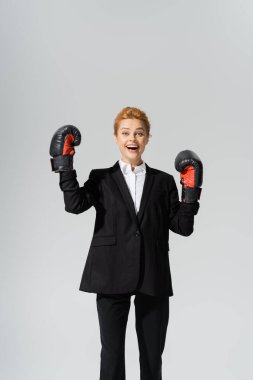 excited redhead businesswoman in black suit and latex gloves rejoicing isolated on grey clipart