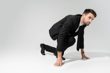 full length of young bearded businessman in stylish formal wear standing in low start position on grey background clipart