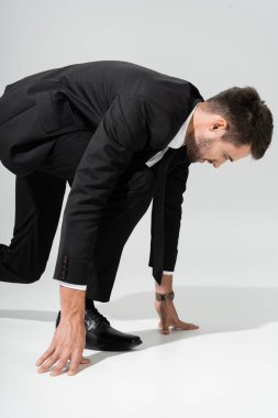 side view of purposeful businessman in black suit standing in low start position on grey background clipart