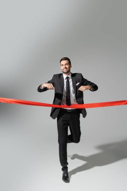 full length of happy businessman in black suit running and crossing red ribbon on grey background clipart