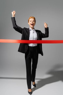 full length of overjoyed businesswoman in black suit crossing finish ribbon on grey background clipart