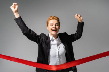 excited redhead businesswoman crossing finish ribbon and showing triumph gesture isolated on grey clipart