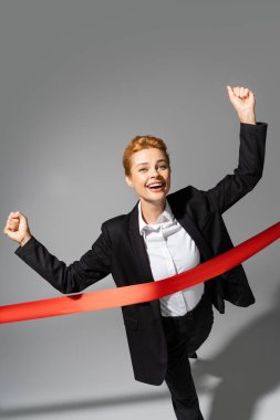 happy businesswoman showing win gesture while crossing finish ribbon on grey background clipart