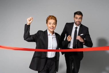 cheerful redhead businesswoman showing win gesture near finish ribbon and business competitor running on grey background clipart