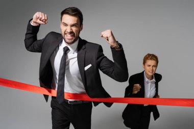 overjoyed businessman in black suit showing win gesture near sad woman running on grey background clipart