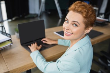 joyful redhead businesswoman typing on blurred laptop with blank screen and smiling at camera in office clipart