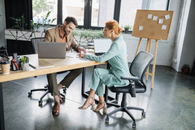 Gülümseyen yönetici, bürodaki kızıl saçlı ve şık iş kadınının yakınındaki belgeleri işaret ediyor.