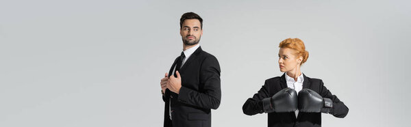 haughty businessman and purposeful businesswoman in boxing gloves looking at each other isolated on grey, banner