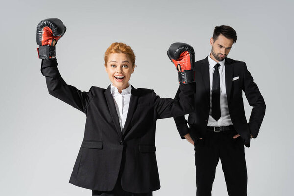 счастливая деловая женщина в боксёрских перчатках, показывающая победный жест вблизи расстроенного делового конкурента, изолированного на сером