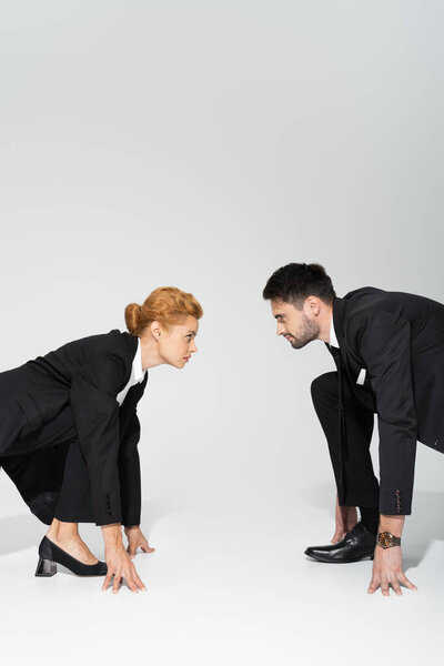 side view of redhead businesswoman and bearded businessman standing in low start position and looking at each other on grey background