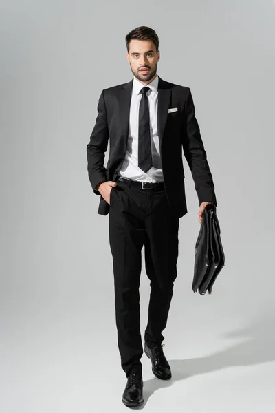灰色の背景を歩きながらポケットに手を入れてブリーフケースを持つスタイリッシュなビジネスマンの完全な長さ — ストック写真