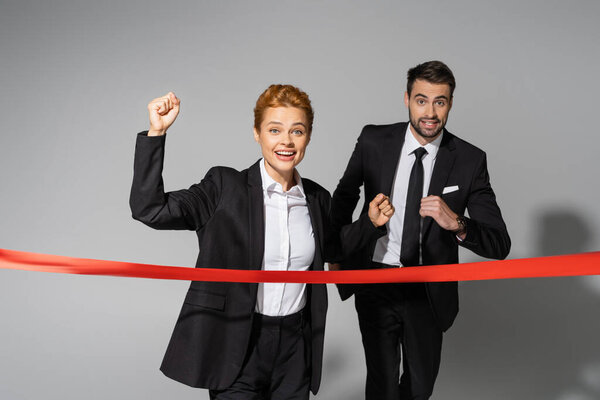 cheerful redhead businesswoman showing win gesture near finish ribbon and business competitor running on grey background