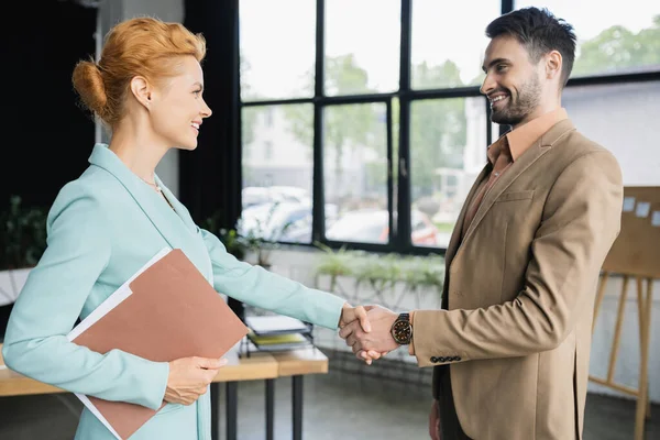 オフィスの若いパートナーと握手するフォルダを持つ幸せなビジネス女性の側の見解 — ストック写真