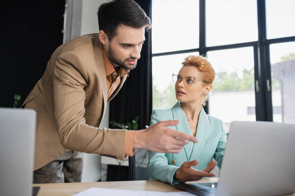bearded businessman pointing at laptop near colleague wearing eyeglasses in office