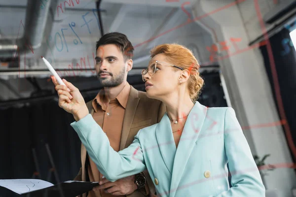 stock image redhead businesswoman pointing at glass board near colleague with folder in office