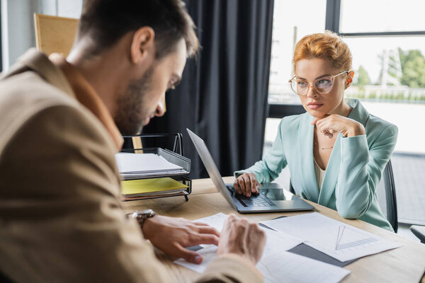 Рыжая бизнесвумен в очках смотрит на размытого менеджера, работающего с бумагами в офисе