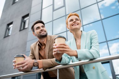 smiling business partners in trendy clothes holding coffee to go and looking away near railing on urban street clipart