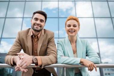 cheerful business partners in trendy blazers smiling at camera near railing on city street clipart