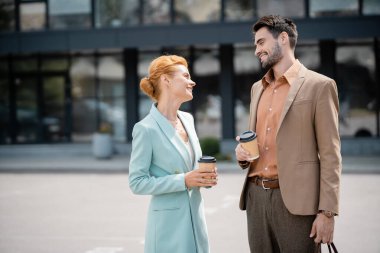 joyful business partners in stylish clothes holding takeaway drinks and talking on city street clipart