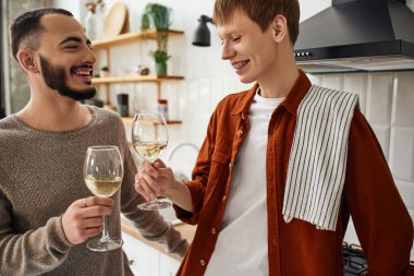 Kızıl saçlı adam mutfakta mutlu sakallı erkek arkadaşıyla şarap bardaklarını tokuşturuyor.