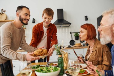 Neşeli eşcinsel çift aile yemeği için ızgara tavuk servis ediyor ve anne babaları mutfakta.