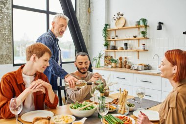 Modern mutfakta genç eşcinsel bir çiftle yemek yerken sakallı bir adam şarap dolduruyor.