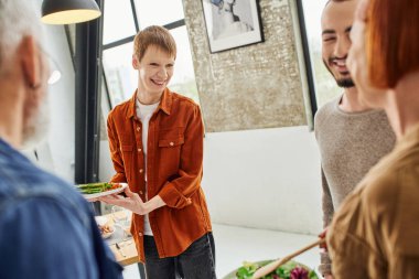 cheerful gay man holding grilled asparagus near blurred boyfriend and parents in kitchen clipart