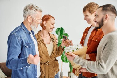 joyful gay couple giving flowers and wine bottle while visiting happy parents at home clipart