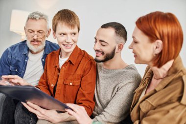 joyful gay man smiling near boyfriend and parents while holding photo album in living room clipart