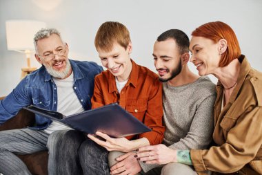 young and happy gay couple looking at photo album while sitting in living room with pleased parents clipart