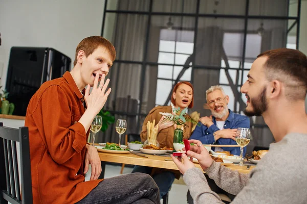 impressed gay man covering mouth with hand near boyfriend making marriage proposal during supper with family at home