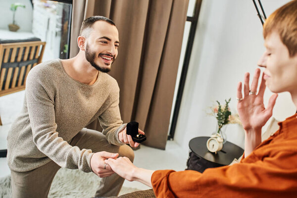 cheerful bearded man holding jewelry box near blurred boyfriend while making marriage proposal at home