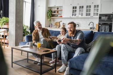 KYIV, UKRAINE - APRIL 19, 2023: Happy gay parents and preteen daughter playing video game on couch at home  clipart