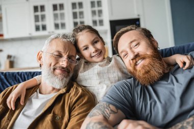 Smiling kid hugging bearded homosexual fathers on couch at home  clipart