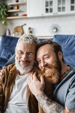 Tattooed and bearded gay man hugging smiling partner at home  clipart