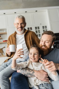 Smiling girl showing peace gesture while taking selfie with homosexual fathers at home  clipart