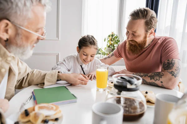 微笑的女孩在同性恋爸爸身边的笔记本上写字 在家里吃早餐 — 图库照片