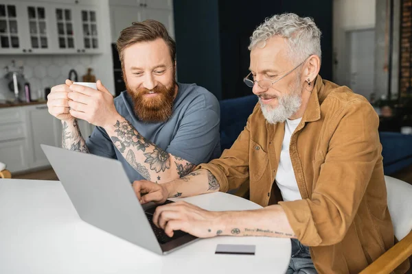 Smiling Gay Man Holding Cup Coffee While Tattooed Partner Using — Stock Photo, Image