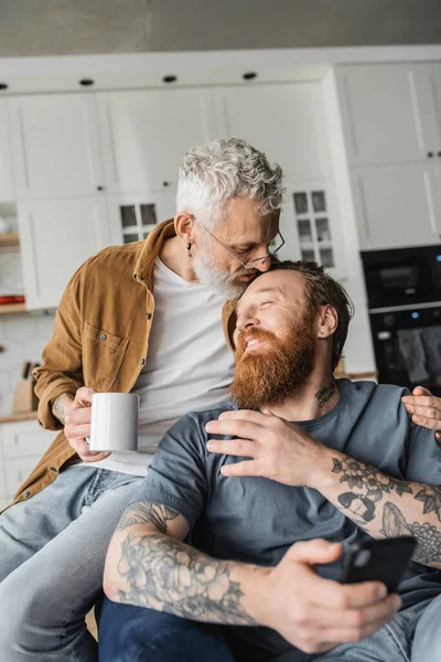 同性男人在家拿着咖啡 用智能手机亲吻纹身的伴侣 — 图库照片