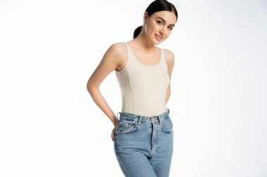 charming and young woman with natural makeup, brunette hair and perfect skin smiling while posing in tank top and denim jeans and looking at camera on white background  clipart