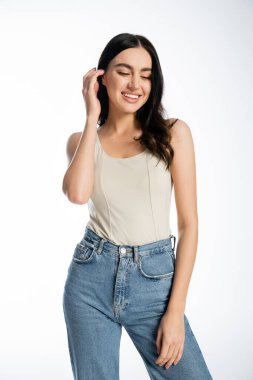 Doğal makyajlı, mükemmel ciltli, gülümseyen, kot pantolon ve kolsuz bluzla poz verirken parlak kumral saçlarını ayarlayan ve beyaz arka plana bakan çekici bir kadın. 