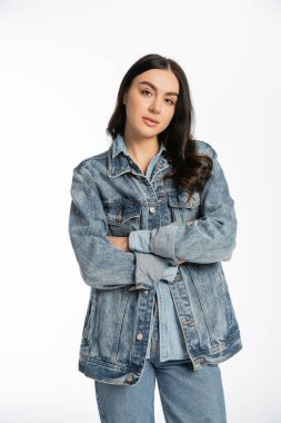 alluring young woman with gorgeous brunette hair posing with folded arms while standing in denim jacket and looking at camera on white background  clipart