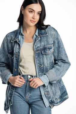 young and fashionable model with brunette hair and natural makeup posing in stylish denim outfit while standing and looking away on white background clipart