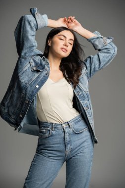 chic young woman with gorgeous hair posing with hands above head and closed eyes while standing in stylish blue jeans and denim jacket on grey background clipart