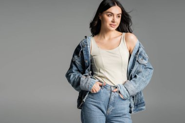 fashionable young woman with brunette hair standing with hands in pockets of blue jeans and posing in stylish denim jacket while smiling isolated on grey background clipart