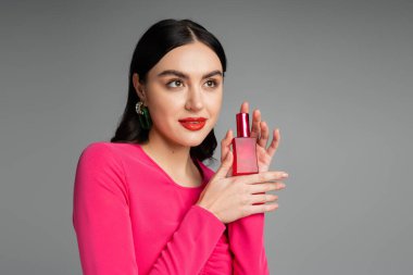Çekici, kumral saçlı, moda küpeli, kırmızı dudaklı ve mor elbiseli, gri arka planda izole edilmiş lüks parfüm şişesi tutan genç bir kadın. 